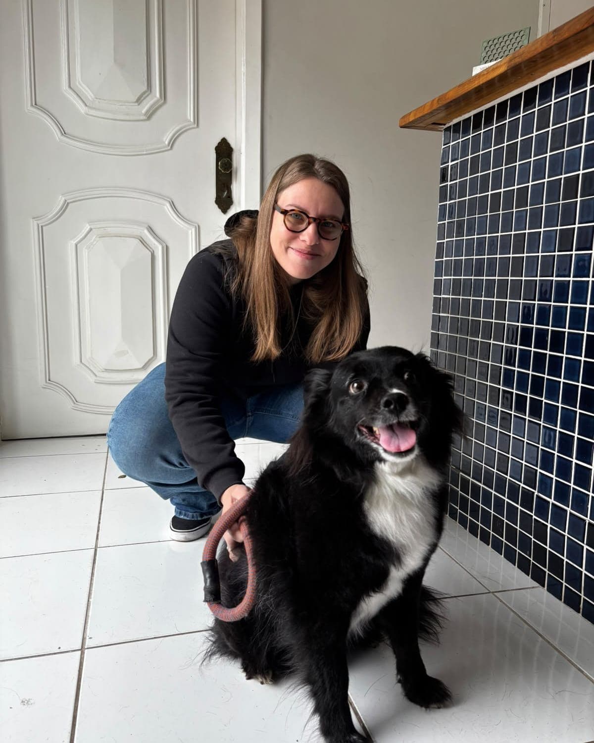 Dra. Mariana Hoffmann agachada ao lado de um cão preto e branco de pelo longo, com a guia na mão, no piso branco da clínica