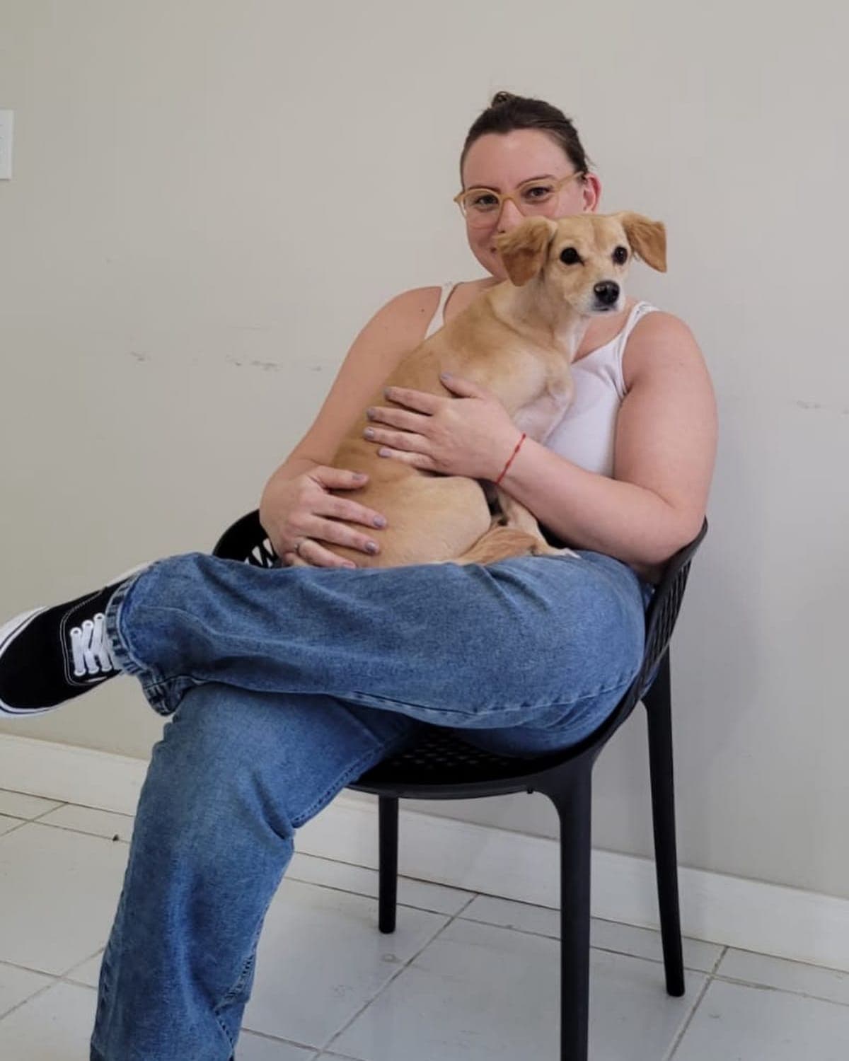 Dra. Mariana Hoffmann sentada numa cadeira preta com um cão caramelo de porte pequeno no colo, olhando pra câmera