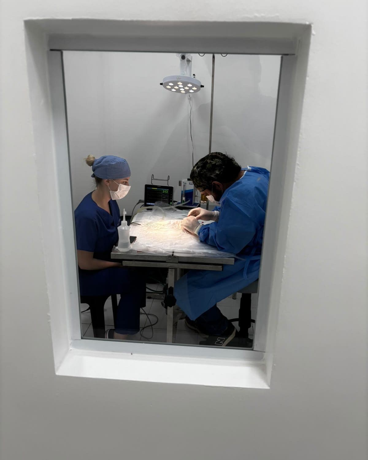 Centro cirúrgico visto pela janela de vidro: dois profissionais paramentados na mesa, com foco cirúrgico aceso e monitor ao fundo