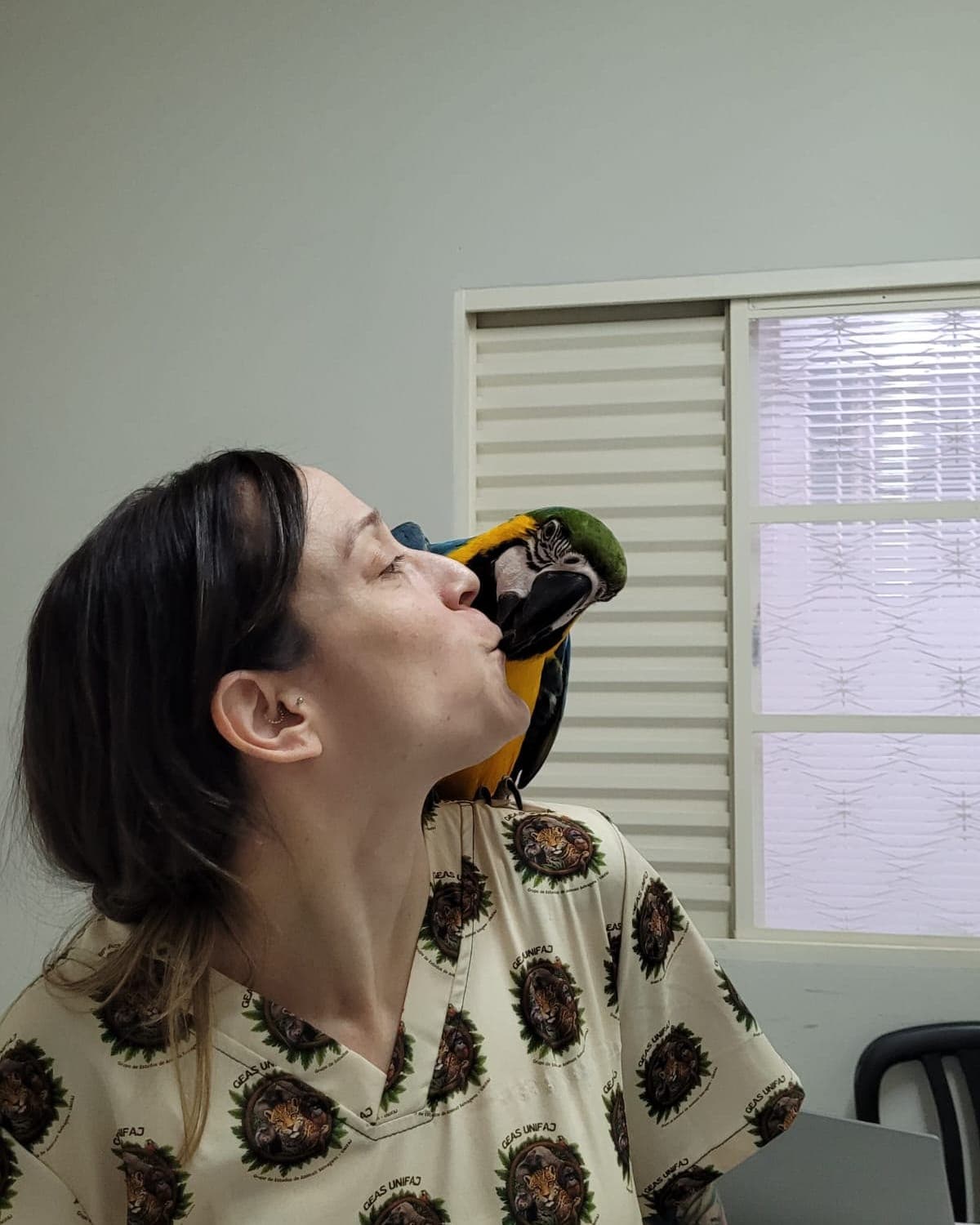 Arara-canindé pousada no ombro da Dra. Mariana Bizzo, encostando o bico no rosto dela, na sala de atendimento